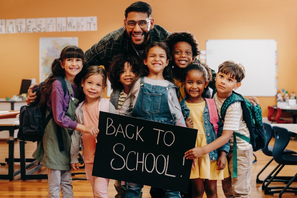 Teacher and his primary school class celebrating coming back to school. Male educator and a group of children smile at the camera while standing in a classroom.